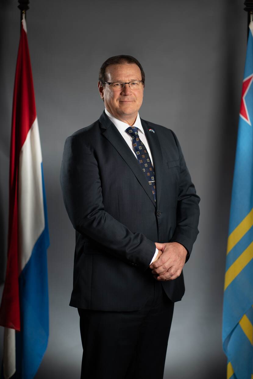 Potret di Gobernador Boekhoudt. Background shinishi scur cu na man robes e bandera Hulandes y na man drechi e bandera di Aruba